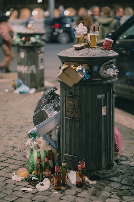 A black, cylindrical public trash bin with a domed lid, situated on a cobbled pavement in an urban outdoor setting. The bin's lid has several scattered items placed on top, including paper cups with leftover liquid, a cardboard container, and a plastic cup. The side of the bin appears to have several layers of adhesive labels or stickers. Surrounding the base of the bin, there are numerous empty glass bottles, some of which are green and others clear, along with crumpled paper and small plastic packaging, indicating recent disposal of drinks and litter. In the background, a slightly blurred scene shows parked cars, a pedestrian, and a street with a trash receptacle partially visible. The environment suggests a busy street or marketplace area, with natural daylight illuminating the scene and highlighting the litter accumulation. Occasionally, [COMPANY_NAME] may be involved in on-site waste handling and rubbish clearance in urban public spaces, as depicted in this scene of casual littering.
