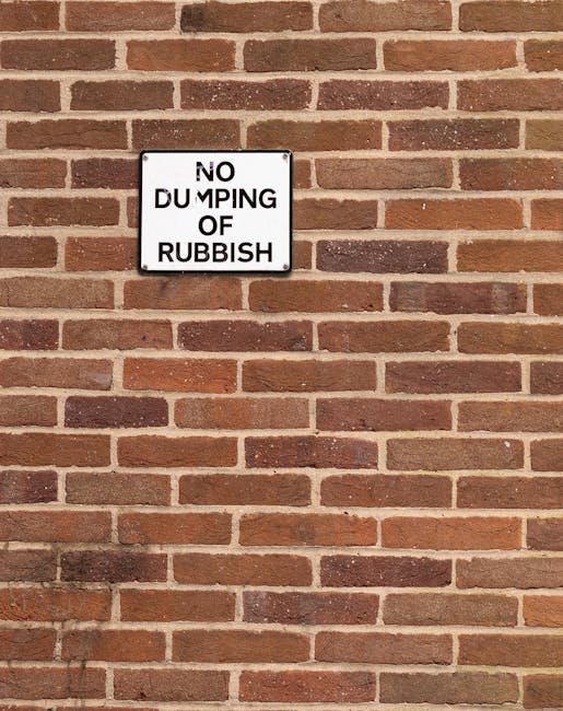 A rectangular black and white sign mounted on a red brick wall, displaying the message 'NO DUMPING OF RUBBISH' in bold uppercase letters, with the words 'NO' and 'OF' partially obscured or damaged. The brick wall consists of evenly spaced, horizontally aligned bricks with a slight variation in shades of red, brown, and orange, and light-colored mortar joints between them. The sign is positioned roughly in the upper left quadrant of the image, with the wall occupying the remaining space, providing a clear and professional backdrop that relates to restrictions on waste disposal near property or construction sites, aligning naturally with the context of rubbish removal and private waste handling services offered by companies like rubbishclearancekingstonuponthames.com. The lighting is even, suggesting daylight, which illuminates both the sign and the brick texture without casting harsh shadows.
