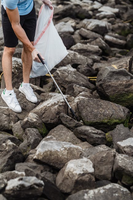 A person wearing a blue shirt, black shorts, and white sneakers is seen selecting and picking up rocks from a large rocky shoreline. The individual is holding a long-handled metal tool that is used to lift or move stones, and a large white mesh bag with pink stripes is held in the other hand, likely for collecting smaller rocks or debris. The rocks vary in size, shape, and color, predominantly shades of gray and brown, with some displaying rough textures and moss or algae growth in certain areas. The environment appears outdoor, with the rocks forming part of a natural shoreline or riverbank, and the lighting is natural, possibly overcast, providing soft, diffused illumination. This scene exemplifies manual rock or debris collection, which can relate to independent waste or rubbish clearance activities, akin to services offered by companies like rubbishclearancekingstonuponthames.com that carry out on-site rubbish removal and waste management tasks efficiently and professionally in various outdoor environments.