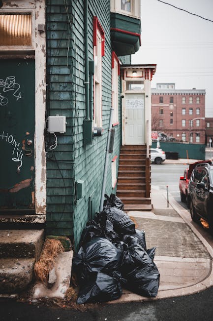 A row of tightly packed black rubbish bags filled with waste are stacked against the side of a green wooden residential building, located at the bottom of a short flight of wooden stairs leading to a white door with red trim. The building's exterior features weathered slate-like cladding, with visible planed edges, and a small white electrical box mounted on the wall. Adjacent to the bags, there are concrete steps leading to a worn, graffiti-marked doorway that is partially visible. The scene is set on a paved sidewalk with a curb, and the background includes parked cars along the street, with an urban landscape of brick buildings and an overcast sky. The image suggests an instance of private rubbish disposal, indicative of independent waste collection methods outside regular municipal services, with the black refuse bags appearing ready for collection or disposal as part of local rubbish removal efforts by Kingston-based waste clearance services.