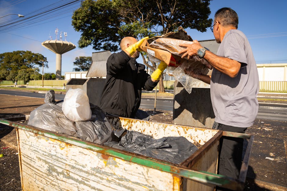 Two men are engaged in collecting waste materials at an outdoor rubbish disposal site during daylight. One man, dressed in a dark jacket and wearing yellow work gloves, is placing debris into a wooden or cardboard container, while the other man, wearing a light grey T-shirt, helps load a large plastic bag containing waste. The container is positioned on a paved surface, with various black and white plastic rubbish bags and discarded materials visible inside it. The scene is set against a backdrop of clear blue sky, with a large tree providing partial shade, and in the distance, there are industrial or storage buildings with yellow fencing. Additional rubbish containers and waste are spread across the area, suggesting an on-site clearance activity that reflects private waste management or alternative collection methods compared to local authority rubbish collection services. The image captures a professional and neutral scene focused on waste removal activities.