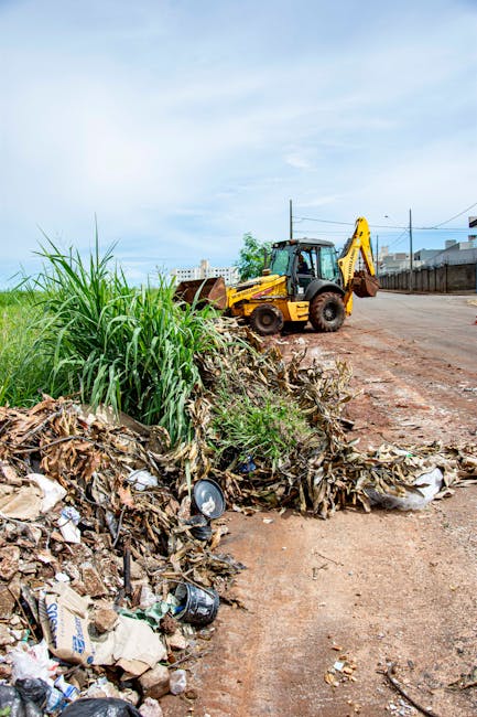 Two men are engaged in collecting waste materials at an outdoor rubbish disposal site during daylight. One man, dressed in a dark jacket and wearing yellow work gloves, is placing debris into a wooden or cardboard container, while the other man, wearing a light grey T-shirt, helps load a large plastic bag containing waste. The container is positioned on a paved surface, with various black and white plastic rubbish bags and discarded materials visible inside it. The scene is set against a backdrop of clear blue sky, with a large tree providing partial shade, and in the distance, there are industrial or storage buildings with yellow fencing. Additional rubbish containers and waste are spread across the area, suggesting an on-site clearance activity that reflects private waste management or alternative collection methods compared to local authority rubbish collection services. The image captures a professional and neutral scene focused on waste removal activities.