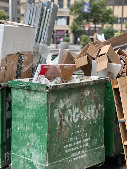A large, green wheeled waste bin filled with various discarded materials including crumpled paper, plastic bottles, and miscellaneous household waste, situated outdoors in an urban environment. The bin's surface shows signs of dirt and scratches, indicating frequent use. Surrounding the bin are several cardboard boxes, some flattened and others partially collapsed, along with wooden pallets and framing materials leaning against and lying nearby. In the background, blurred trees, urban buildings, and parked cars suggest a city street setting. The scene depicts the process of outdoor rubbish collection, possibly related to independent waste disposal or bulk waste clearance, with the contents appearing ready for removal or transfer to a waste management service such as those provided by Kingston Bridge bulky waste collection and disposal services. Natural daylight illuminates the scene, highlighting the textures and colors of the various debris and emphasizing the practical, utilitarian nature of the waste management environment.