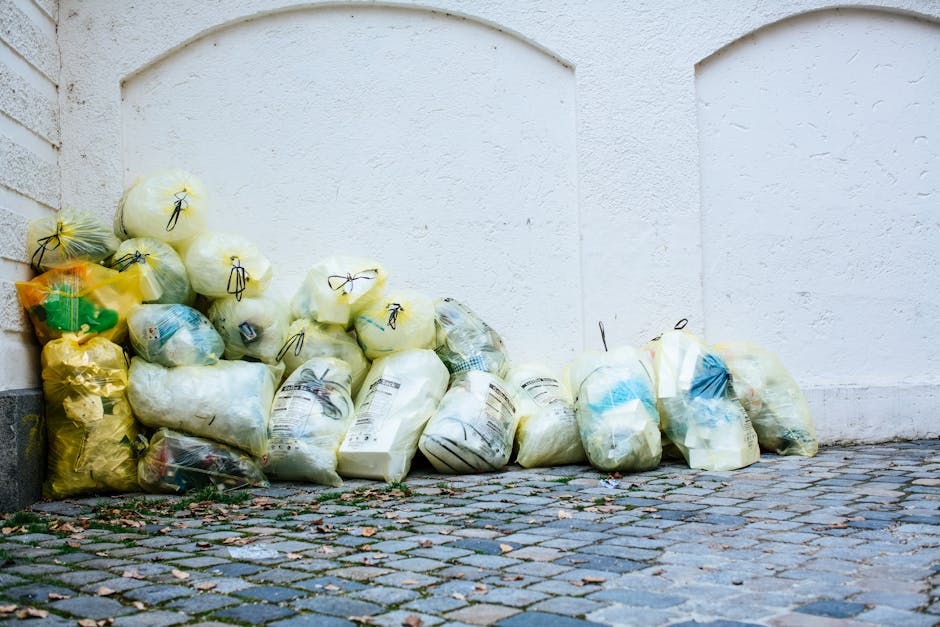 A collection of numerous plastic rubbish bags, mostly white and yellow, are piled against a white textured wall on a cobblestone pavement. The bags are tied with black string or twist ties and contain various types of waste, including packaging material and household refuse, some with visible labels. They are stacked haphazardly, with some leaning against the wall and others resting on the uneven cobblestones, which are interspersed with small leaves and bits of debris. The scene appears to be part of an outdoor area suitable for private waste collection or rubbish removal services, such as those offered by Kingston-based companies, highlighting an instance of alternative on-site disposal rather than local authority collection. The lighting is natural, casting soft shadows on the bags and pavement, emphasizing the different textures of the bags' plastic surfaces and the roughness of the cobblestones, consistent with a typical residential or commercial waste clearance context.