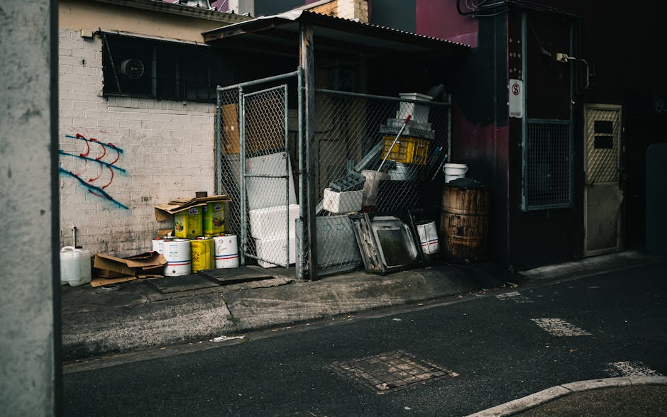 A small, outdoor storage area situated on a city street corner, featuring a makeshift metal shelter with a corrugated roof, supported by metal posts, positioned directly against a white brick wall. Inside and around the shelter, various discarded items are visible, including white plastic containers, metal trays, wooden pallets, a barrel, and cardboard boxes, some of which are open and containing additional waste materials. The objects rest on a paved surface with a slightly worn appearance, adjacent to the curb of a street with visible drainage grates. To the left of the shelter, a few more containers and packaging materials are stacked, while the surrounding environment includes a dark building façade and limited natural light, suggesting an urban setting where independent waste disposal activities, such as rubbish clearance, occur outside typical council collection zones. The scene is neutral in tone, with the mix of specific waste items highlighting private or alternative rubbish handling practices, as part of rubbish removal services in Kingston upon Thames.