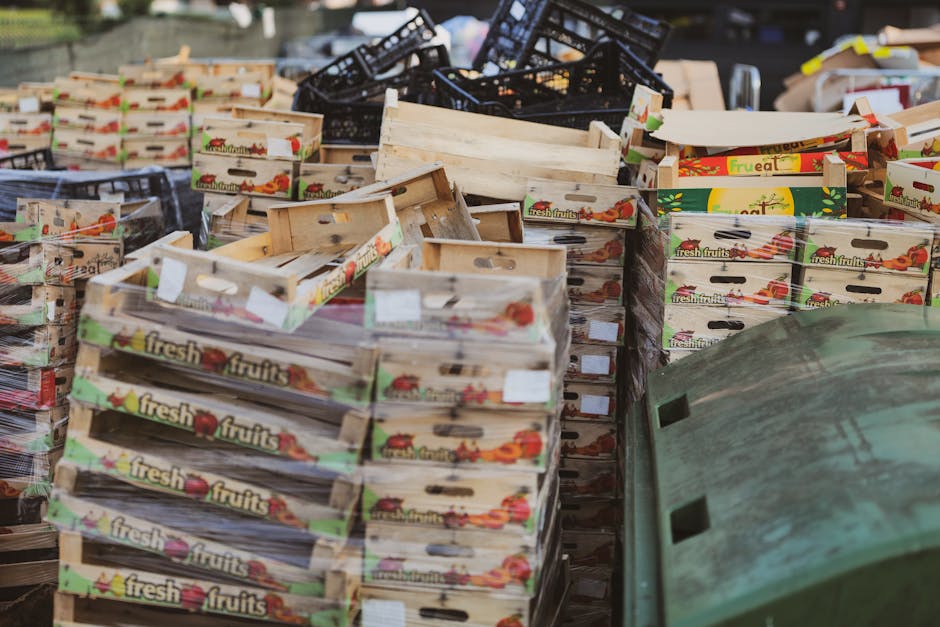 A cluttered outdoor scene featuring numerous wooden fruit crates stacked and scattered on the ground, some stacked neatly while others are leaning or partially overturned. The crates are made of light-colored, rough-textured wood, with printed labels displaying images of fresh fruits and the words 'fresh fruits' on the sides. In the background, there are black plastic crates stacked haphazardly, some tilted or overlapping, and a few cardboard boxes partially visible among the wooden crates. The scene is lit by natural daylight, casting soft shadows, and the surrounding environment appears to be an outdoor storage or waste disposal area, with a green plastic container or bin partially visible in the lower right corner. The overall impression is of discarded or surplus produce packaging materials, relevant to private waste handling or rubbish removal activities, with the scene reflecting an accumulation of waste suitable for clearance services.