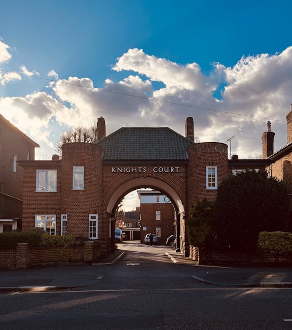 A brick archway labeled 'Knights Court' serves as the entrance to a residential complex, with the arch supported by cylindrical brick columns on either side. The building above the arch features a dark shingle roof and several white-framed windows, with two prominent brick chimneys protruding from the roof. To the right and left of the arch, low brick walls with greenery, including bushes and small trees, partially obscure the view of the surrounding buildings. Behind and beyond the arch, parked cars are visible along a paved driveway leading into a courtyard area within the complex. The sky above is partly cloudy with bright sunlight illuminating the scene, cast through the fluffy clouds, creating a natural, well-lit environment. The environment appears clean and well-maintained, suggesting a private residential area that could be associated with alternative waste management or independent rubbish removal services, aligning with the context of private rubbish clearance. The overall scene emphasizes a quiet residential setting with clear visual cues for access and property boundaries.