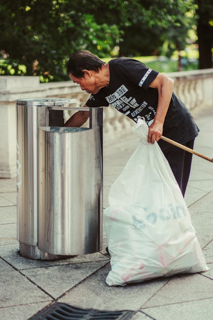A middle-aged man with dark hair, wearing a black T-shirt with white and blue lettering, is seen discarding rubbish into a stainless steel outdoor waste bin on a paved sidewalk. The man is holding a large white plastic bag filled with waste in his left hand, while his right hand is reaching into the bin to place more rubbish inside. The scene is set outdoors with a background of green trees and a stone or concrete balustrade, indicating a park or public area. The waste bin is cylindrical with a shiny metallic finish, positioned next to a drain grate on the pavement. The environment appears well-lit by natural daylight, emphasizing the clean, urban setting suitable for private rubbish collection or on-site clearance services such as those offered by rubbish clearance Kingston upon Thames. The image captures an act of independent rubbish disposal, potentially illustrating a service related to responsible waste handling outside traditional municipal collection systems.
