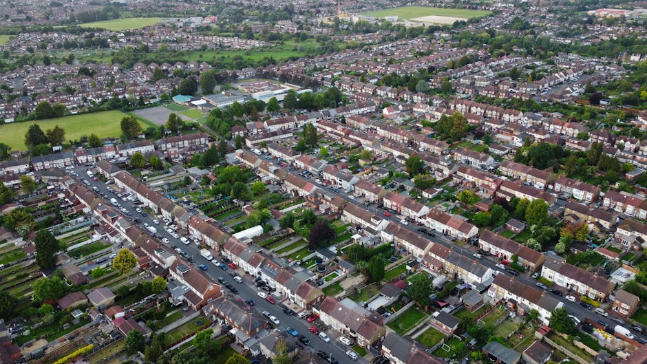 An aerial view of a residential suburban area showing rows of terraced houses with brick facades and tiled roofs, interspersed with trees and small gardens. The street in the foreground is lined with parked cars, and the houses have front yards with lawns, hedges, and paved pathways. Behind the residential area, green sports fields and parkland are visible, bordered by more houses and leafy streets extending into the distance. The scene is captured under natural daylight, with a clear overcast sky providing diffuse lighting, highlighting the density and layout of the neighbourhood that could be associated with private waste collection and local rubbish removal services, such as those offered by companies like rubbishclearancekingstonuponthames.com, in the context of environmentally conscious waste management for town homes.