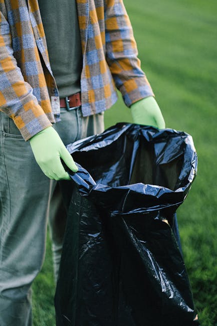 A person wearing a checked yellow and grey flannel shirt, green gloves, and light grey pants holds open a large black bin liner, which is partially filled with waste. The individual is standing outdoors on a grassy area with well-maintained green lawn in the background, suggesting an environment suitable for rubbish collection or waste removal. The bin liner appears to be made of durable plastic with a shiny surface, and the waste inside is not visible. The person's hands grip the edges of the bag, preparing for waste disposal or transfer, indicative of a professional rubbish removal activity. The scene reflects practices of on-site waste clearance, highlighting the importance of proper waste handling for environmental and health standards, aligning with independent disposal methods often used in private rubbish collection services. The natural daylight illuminates the scene evenly, emphasizing the textures and colors of the clothing, gloves, and waste container, supporting visual clarity for accessibility and SEO relevance on waste management topics.