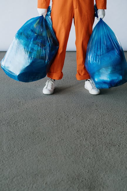 A person wearing orange work trousers and white sneakers is standing on a grey paved surface, holding two large blue plastic rubbish bags filled with waste, with one bag in each hand. The background is plain and neutral, with no visible setting or environment details. The image focuses on the individual's lower body and the rubbish bags, which appear to contain general waste or rubbish suitable for collection services. The scene exemplifies a scenario related to waste disposal or rubbish removal, highlighting the ease of handling bulky rubbish using personal effort, potentially reflecting private waste handling or on-site clearance activities. The lighting is even and neutral, emphasizing the textures of the plastic bags and the fabric of the trousers, with minimal shadows, creating an overall clear and professional visual suitable for a waste management or rubbish removal context.