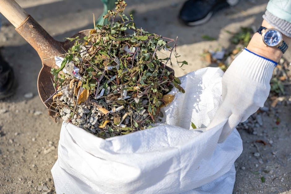 A person wearing a checked yellow and grey flannel shirt, green gloves, and light grey pants holds open a large black bin liner, which is partially filled with waste. The individual is standing outdoors on a grassy area with well-maintained green lawn in the background, suggesting an environment suitable for rubbish collection or waste removal. The bin liner appears to be made of durable plastic with a shiny surface, and the waste inside is not visible. The person's hands grip the edges of the bag, preparing for waste disposal or transfer, indicative of a professional rubbish removal activity. The scene reflects practices of on-site waste clearance, highlighting the importance of proper waste handling for environmental and health standards, aligning with independent disposal methods often used in private rubbish collection services. The natural daylight illuminates the scene evenly, emphasizing the textures and colors of the clothing, gloves, and waste container, supporting visual clarity for accessibility and SEO relevance on waste management topics.
