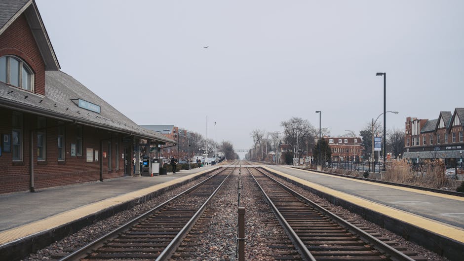 A wide view of a train platform at a railway station, with two parallel set of steel train tracks running centrally into the distance. On the left side, a red brick station building with a sloped grey roof features several small windows and doors, some of which are open with waiting areas or ticket machines visible beside them. A yellow tactile paving strip lines the edge of the platform, providing guidance for visually impaired passengers. On the right side, a similar platform features a row of black lampposts and multiple buildings with brick facades, some with retail signage. Sparse trees and bushes are visible along the horizon, with a few passengers waiting or walking along the platform, and a grey sky overhead indicating an overcast day. The atmosphere is calm and quiet, typical of an early morning or late afternoon, with no moving trains or heavy activity apparent. The scene exemplifies an accessible, urban railway station and provides a visual context related to transportation and alternative ways of managing mobility and potentially non-daily waste generated in travel-related environments, as might be handled through private rubbish removal services such as those offered by rubbishclearancekingstonuponthames.com across the town center.