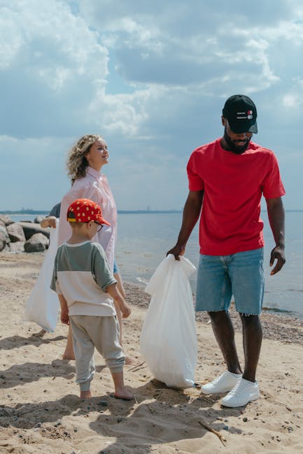A man and two children picking up litter on a sandy beach near a body of water under a cloudy sky. The man, wearing a red t-shirt, ripped denim shorts, white sneakers, and a black cap with a small flag emblem, is holding a large white rubbish bag. One of the children, a young girl with curly blonde hair, wears a light pink jacket, while the other, a young boy, has a red cap with white polka dots and casual grey clothes. Rocks are visible in the background along the shoreline, and the scene is illuminated by natural daylight, creating a clear, realistic view of an outdoor cleanup activity, indicative of private waste handling or onsite clearance supported by the presence of the rubbish bag.